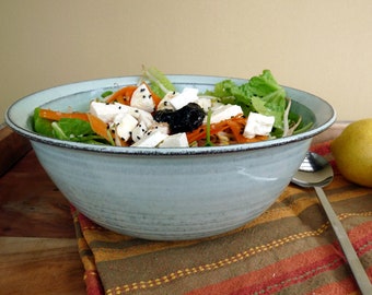 May include: A light blue ceramic bowl filled with a salad of lettuce, carrots, feta cheese, and black sesame seeds. The bowl is sitting on a striped placemat with two lemons and a silver spoon in the background.