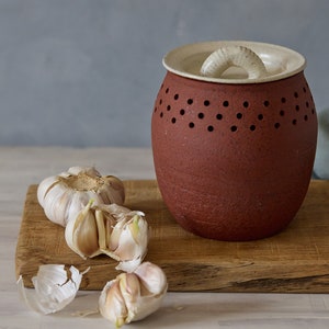 May include: A red ceramic garlic keeper with a white lid and a handle. The keeper has small holes for ventilation. There are four garlic cloves on a wooden chopping board next to the keeper.