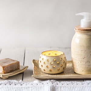 Puede incluir: Un conjunto de tres accesorios de baño de cerámica en color crema con un acabado moteado. El conjunto incluye un jabonera, un portavelas y una botella de bomba con un tapón de corcho.