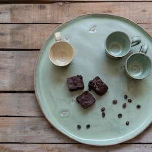 Puede incluir: Una bandeja de servir de cerámica verde claro con cuatro cuadrados de chocolate y tres tazas de cerámica. La bandeja tiene un aspecto rústico y hecho a mano y está decorada con diseños de hojas.