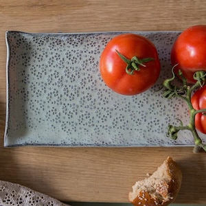May include: A rectangular, light blue ceramic serving dish with a floral pattern holds three ripe red tomatoes on a wooden surface. A piece of bread and a lace cloth are also present.