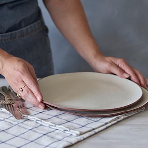 Puede incluir: Dos platos de cerámica blanquecina con borde marrón rojizo apilados sobre un paño a cuadros. Cubiertos, incluyendo tenedores y cuchillos, están dispuestos a la izquierda. Se están manipulando los platos.