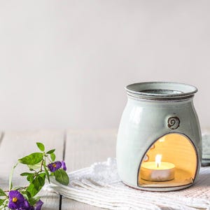 May include: A pale blue ceramic oil diffuser with a curved opening for a tea light. The diffuser features a spiral design and sits on a white cloth. Purple flowers and green leaves are in the foreground.