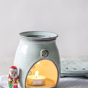 Puede incluir: Un quemador de aceite de cerámica blanca con una pequeña vela de té en su interior. El quemador tiene un diseño decorativo de espiral en la parte delantera y un recorte para la vela. Una pequeña figura de Papá Noel se encuentra en primer plano.