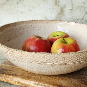 May include: A white ceramic bowl with a speckled glaze sits on a wooden surface. Three red apples are inside the bowl.