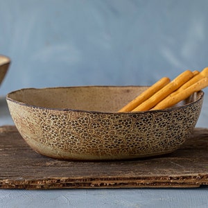 May include: A handmade ceramic bowl with a textured floral design and a dark brown rim. The bowl holds several breadsticks. The bowl is placed on a piece of rustic, weathered wood. The background is a muted blue.