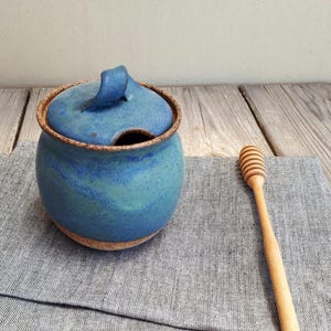 May include: A handmade blue ceramic honey pot with a lid and a built-in spoon rest. The pot has a rustic, textured appearance with a brown rim and base. A wooden honey dipper rests beside the pot on a gray cloth.
