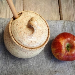 May include: A beige ceramic honey pot with a lid and a wooden honey dipper sticking out of the lid. The pot has a speckled glaze and a knob on the lid. A red apple sits next to the pot on a gray fabric surface.