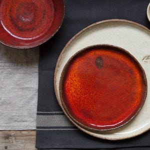 Peut inclure: Vue aérienne de vaisselle en céramique faite à la main. Un bol et une assiette rouge foncé sont visibles, ainsi qu'une assiette crème et un petit bol. Les pièces sont posées sur un tissu gris foncé et une surface en lin gris clair.