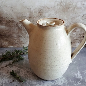 Pode incluir: Uma chaleira de cerâmica branca com um esmalte salpicado e uma tampa. A chaleira tem um corpo arredondado e uma alça lateral.