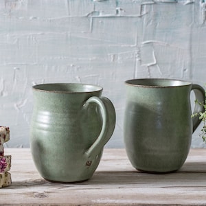 May include: Two green ceramic mugs with rounded handles on a white wooden surface. A stack of three pink and white candy squares is in the foreground.