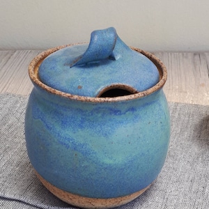 Puede incluir: Un azucarero de cerámica azul y verde con tapa y asa. El cuenco tiene un esmalte moteado y un borde marrón.