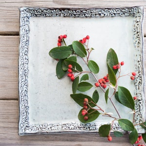 Puede incluir: Un plato cuadrado de cerámica blanca con un borde decorativo, que presenta una rama de hojas verdes con bayas rojas.