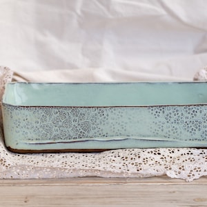 Puede incluir: Un plato rectangular de cerámica azul claro con un patrón decorativo alrededor del borde. El plato está sobre una puntilla blanca.