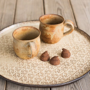 Może przedstawiać: Ceramiczna taca do serwowania z dwoma kubkami i trzema czekoladowymi trufelami. Kubki i taca mają nakrapiany, ziemisty odcień. Taca ma dekoracyjny wzór w kropki. Kubki mają uchwyty.