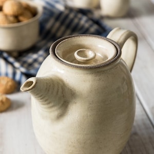 Puede incluir: Una tetera de cerámica de color crema con tapa y borde marrón. La tetera está sobre una superficie de madera blanca.