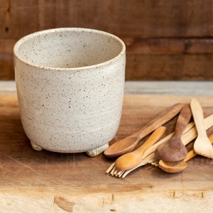 Puede incluir: Una taza de cerámica blanca moteada con forma redondeada y pequeños pies, junto a una colección de cucharas de madera de varios tamaños y tonos. Los artículos están dispuestos sobre una superficie de madera.