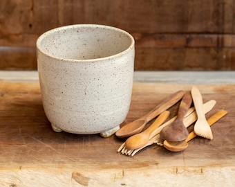 May include: A speckled white ceramic cup with a rounded shape and small feet, next to a collection of wooden spoons of various sizes and shades. The items are arranged on a wooden surface.