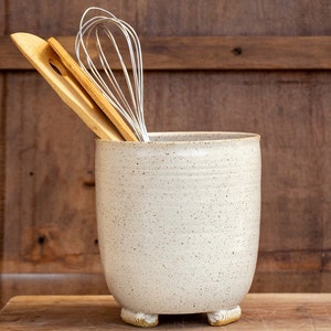 Op de afbeelding: Een witte keramische bestekhouder met een gespikkelde glazuur en drie kleine voetjes. De houder is gevuld met een houten lepel en een metalen garde.
