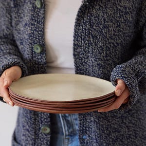 May include: A stack of five ceramic plates. The plates have a light beige center and a dark brown rim. The plates are held by a person wearing a blue knit cardigan.
