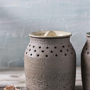 Könnte beinhalten: Ein rustikaler, grauer Knoblauchtopf aus Keramik mit Deckel und kleinen Belüftungslöchern. Der Topf steht auf einer Holzoberfläche, Knoblauchzehen liegen daneben. Schlichtes, funktionales Design für die Küchenaufbewahrung.
