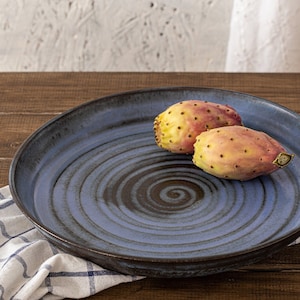 May include: Two prickly pear cactus fruits on a blue and brown ceramic plate with a spiral design. The plate is on a wooden table with a white and blue checkered cloth.