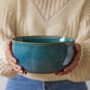 May include: A large, teal ceramic bowl with a brown rim. The bowl is being held by a person's hands.