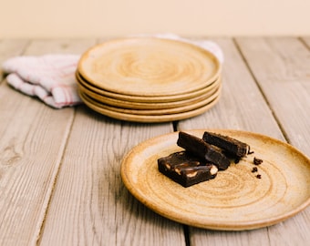 May include: A close-up of a stack of four brown ceramic plates with a single plate on a wooden table. A small pile of chocolate brownies sits on the plate in the foreground.