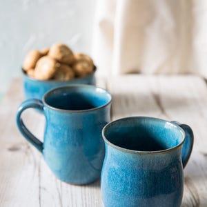 Puede incluir: Dos tazas de cerámica azul con acabado esmaltado sobre una mesa de madera blanca. Las tazas tienen una forma redondeada y un asa en el lateral.