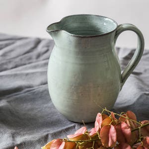 May include: A light blue ceramic jug with a wide mouth and a handle. The jug is sitting on a grey linen tablecloth with a sprig of pink flowers.