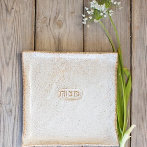 May include: A square, off-white ceramic plate with the Hebrew word "Matzah" embossed in the center. The plate has a textured surface and a slightly rough, brown edge. A sprig of white flowers and green leaves rests beside the plate on a wooden surface.