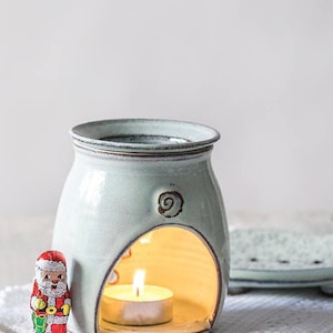Può includere: Un diffusore di oli essenziali in ceramica azzurra con un'apertura curva, una candela accesa all'interno e un motivo a spirale. Una piccola statuina di Babbo Natale è sulla sinistra. Un piatto in ceramica abbinato è sullo sfondo.