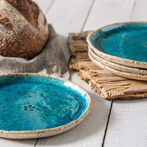 May include: A set of four teal blue ceramic plates with a speckled glaze and a raised design in the center. The plates are stacked on a piece of wood.