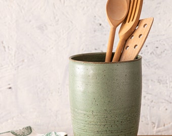 May include: A green ceramic utensil holder with a speckled finish, holding wooden kitchen utensils, including a spoon, fork, and spatula.