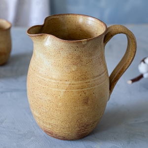 May include: A speckled, light brown ceramic pitcher with a wide mouth and a handle. The pitcher has a rustic, handmade look.