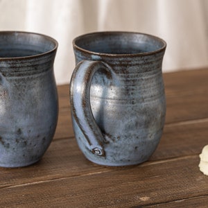 May include: Two blue and brown ceramic mugs with rounded handles on a wooden surface.  A stack of four white donut-shaped cookies is on the right side of the image.