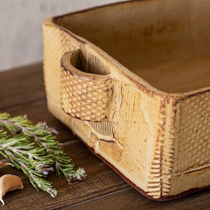May include: A rectangular ceramic baking dish with a textured surface and a handle on one side. The dish is a light brown color and has a slightly reddish brown rim.