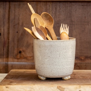 May include: A speckled, light grey ceramic utensil holder containing wooden spoons, forks, and spatulas. The holder has a rounded shape and sits on three small feet. The wooden utensils vary in size and shape.