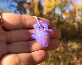 Puede incluir: Un collar con colgante en forma de corazón translúcido, en tonos lavanda y rosa. El colgante está suspendido de una delicada cadena de plata. El collar se sostiene en una mano sobre un fondo borroso de árboles y cielo azul.