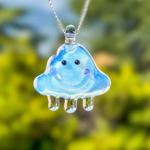 Peut inclure: Un pendentif en verre bleu en forme de nuage avec un visage souriant et trois pieds en forme de larme. Le pendentif est suspendu à une chaîne argentée.