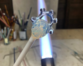 Puede incluir: Un colgante de vidrio transparente con un diseño de estrella está siendo calentado con un soplete. El colgante está unido a una varilla de vidrio blanco.