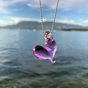 May include: A delicate silver chain necklace with a light purple glass heart pendant. The heart is slightly asymmetrical and has a translucent quality.