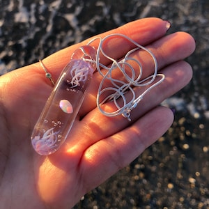 May include: A clear glass pendant necklace with a silver chain. The pendant contains a jellyfish design with white tentacles and a pearlescent opal-like stone. The necklace is displayed on a hand.