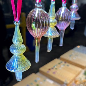 Könnte beinhalten: Eine Reihe schillernder Glasornamente mit rosa Bändern zum Aufhängen. Die Ornamente haben verschiedene Formen, darunter Tropfen, Kugeln und Zierknöpfe. Das Glas reflektiert Farben wie Gelb, Blau und Rosa. Im Vordergrund sind hölzerne Präsentationsbretter zu sehen.