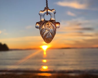 Puede incluir: Un colgante de corazón de vidrio transparente con detalles intrincados cuelga sobre un fondo de puesta de sol sobre una masa de agua.