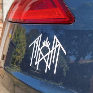 May include: A white vinyl decal featuring a stylised geometric emblem with sharp, angular lines and a central sunburst motif, applied to the dark blue glossy surface of a vehicle near the red taillight assembly.
