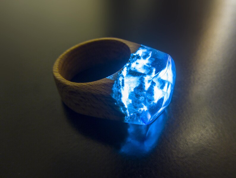 May include: A wooden ring with a blue resin inlay. The resin is translucent and has a swirling pattern. The ring is a unique and stylish piece of jewelry.