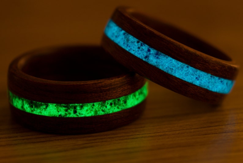 Puede incluir: Dos anillos de madera con una incrustaci&oacute;n de resina verde y azul que brilla. Los anillos est&aacute;n hechos de madera oscura y tienen un acabado liso.