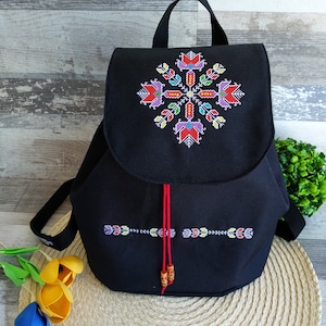 Op de afbeelding: Zwarte canvas rugzak met een kleurrijk geborduurd ontwerp. Het ontwerp heeft een geometrisch patroon met rode, gele, groene, blauwe en paarse accenten.