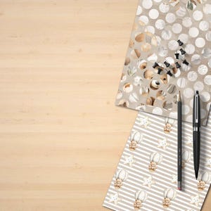 Puede incluir: Un escritorio de madera con art&iacute;culos de papeler&iacute;a y papel estampado. El papel presenta osos de peluche, estrellas y globos aerost&aacute;ticos. Un l&aacute;piz negro, un bol&iacute;grafo negro y chinchetas negras tambi&eacute;n est&aacute;n en el escritorio.